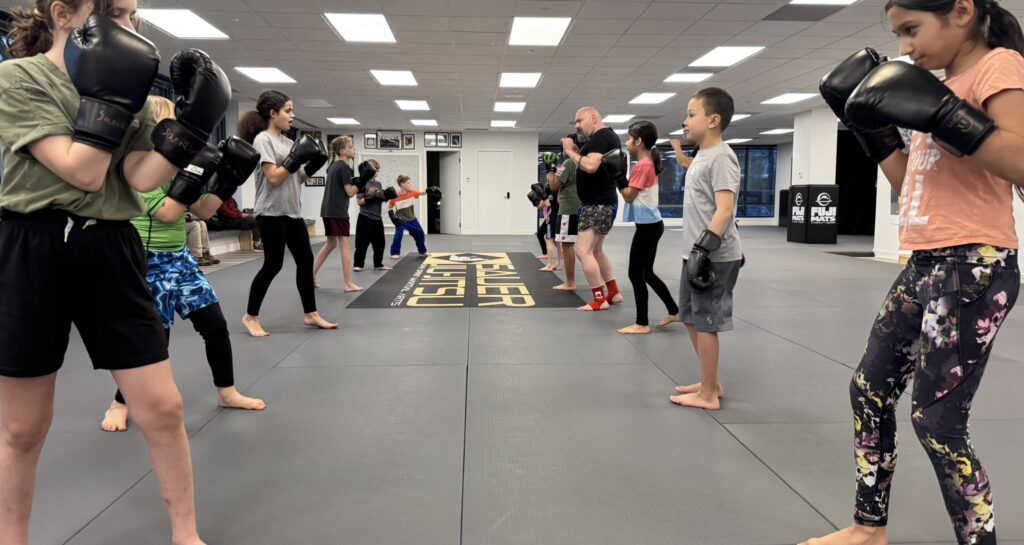 Coach Kevin Teaching Youth Muay Thai Class in Herndon, VA - Pedro Sauer Academy - Pedro Sauer Academy Coach Kevin instructing students during a Youth Muay Thai class at Pedro Sauer Academy in Herndon, Virginia, as children wearing boxing gloves prepare for partner drills.
