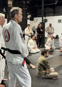 Aaron - Pedro Sauer Academy - Pedro Sauer Academy Man in a white gi with a black belt stands at the front while others kneel on mats in a training dojo.