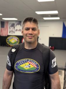 IMG_0880 - Pedro Sauer Academy - Pedro Sauer Academy Smiling man in a black rash guard with a backpack in a martial arts gym setting.