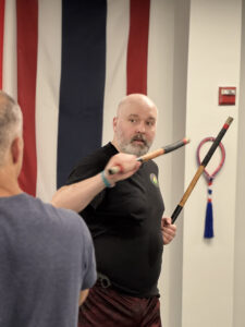 IMG_1256 - Pedro Sauer Academy - Pedro Sauer Academy Bearded man in a black shirt holding a wooden staff indoors, with striped wall panels behind him.
