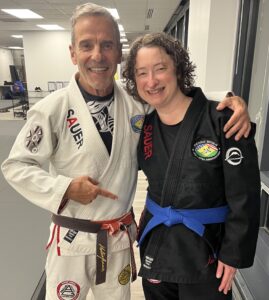 Mary - Pedro Sauer Academy - Pedro Sauer Academy Two smiling martial artists pose together in a dojo; man in a white gi with brown belt and patches, woman in a black gi with blue belt, arm around each other, indoors.