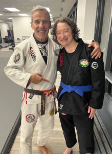 Mary (TBD) - Pedro Sauer Academy - Pedro Sauer Academy Two smiling martial artists pose in a dojo: man in a white gi with patches and a brown belt, woman in a black gi with a blue belt.