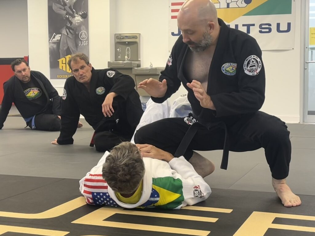 Jiu-jitsu instructor demonstrates a ground grappling move on a student while three observers watch in a dojo gymnasium.