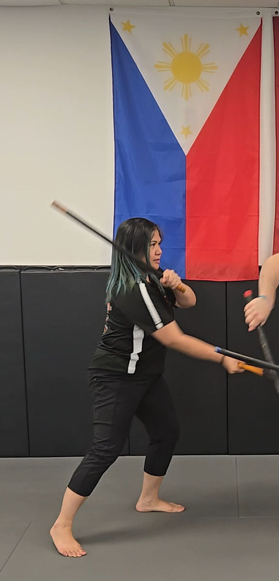 kim-coach - Pedro Sauer Academy of Self-Defense - Pedro Sauer Academy of Self-Defense Coach Kim practices Kali with double sticks in front of the Philippine flag at Pedro Sauer Academy.