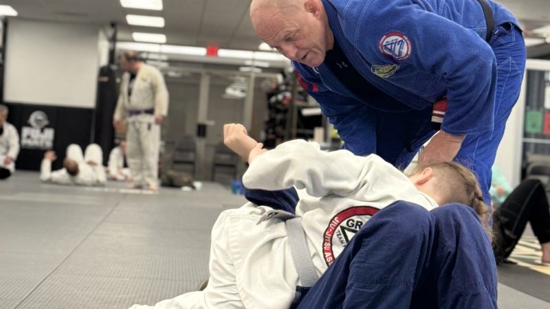 Coach T., a first-degree Brazilian Jiu-Jitsu black belt in a blue gi, assists two students during a youth BJJ class at Pedro Sauer Academy in Herndon, Virginia.