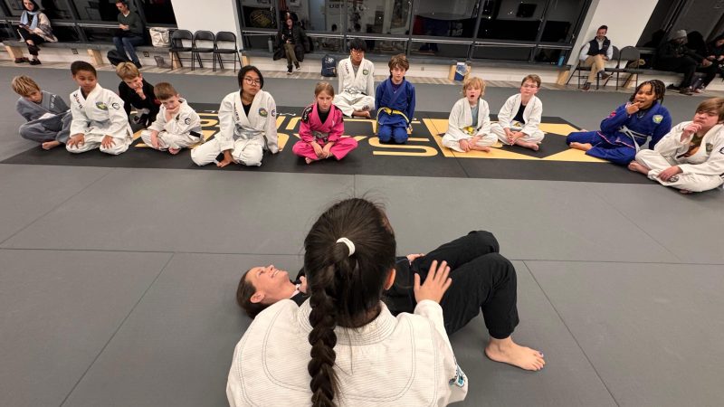 Youth Brazilian Jiu-Jitsu class at Pedro Sauer Academy with children watching an instructor demonstrate a ground technique